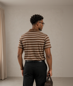 A man viewed from behind wearing a brown and beige horizontally striped short-sleeve t-shirt, black trousers, and holding a brown leather briefcase against a neutral studio backdrop.