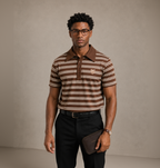 A man wearing a brown and beige horizontally striped polo shirt with a small embroidered fox logo on the chest, paired with black trousers and holding a dark brown leather clutch against a neutral studio backdrop.